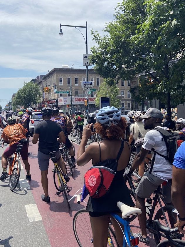 Cyclists in helmets riding together down a Brooklyn bike lane past shops and trees