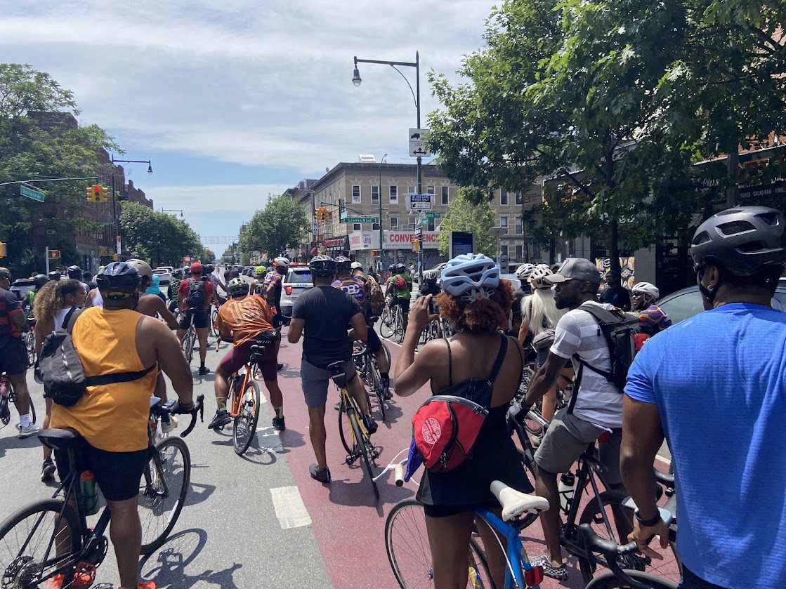 A large group of cyclists in helmets riding together along a red bike lane in Brooklyn on a sunny day