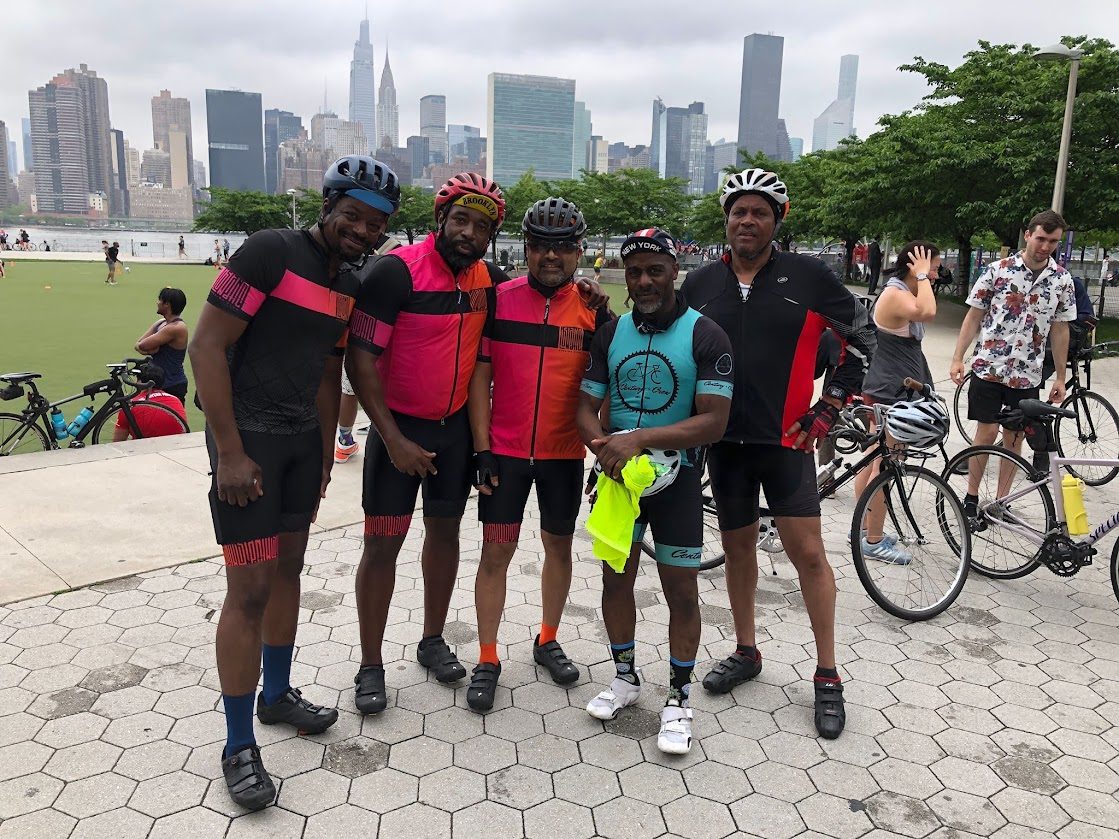 Five cyclists in bright club jerseys standing together on a waterfront plaza with the Manhattan skyline behind them