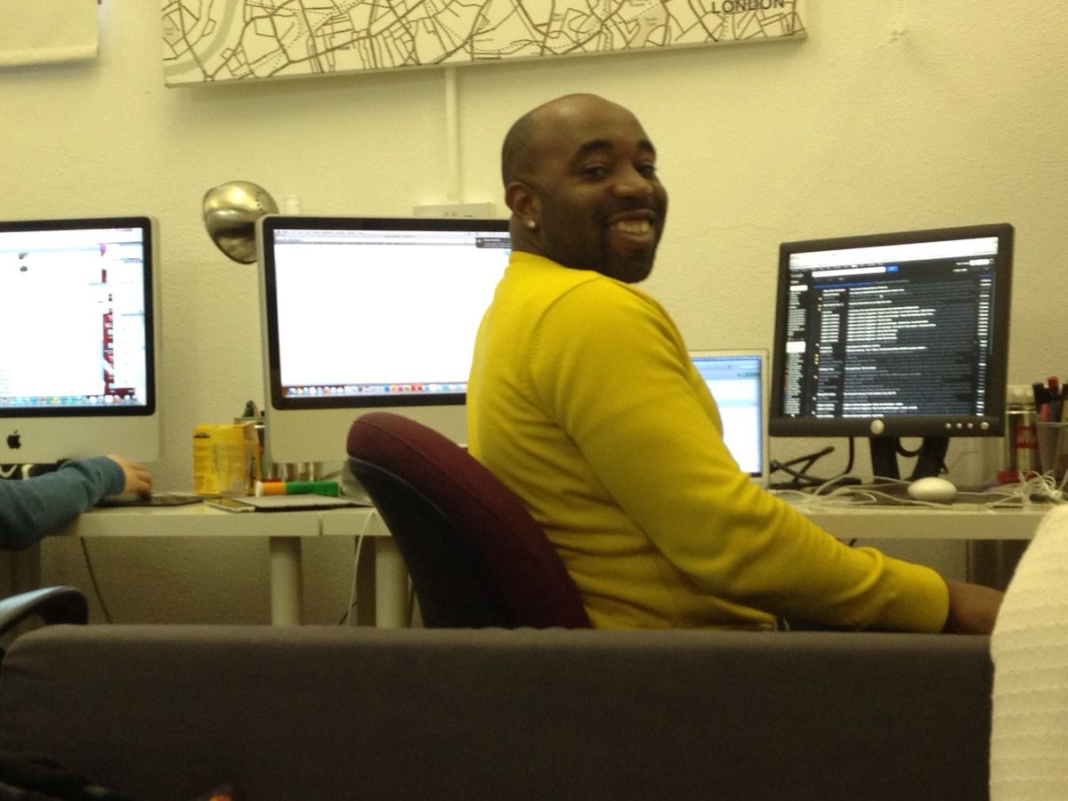 Kenny Akpan smiling at his desk in a London office while a colleague works at the next computer