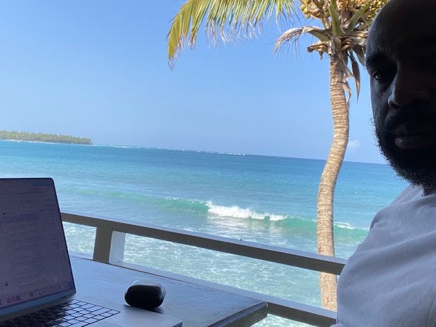Kenny Akpan working on a laptop at a table overlooking the sea, with a palm tree and breaking waves