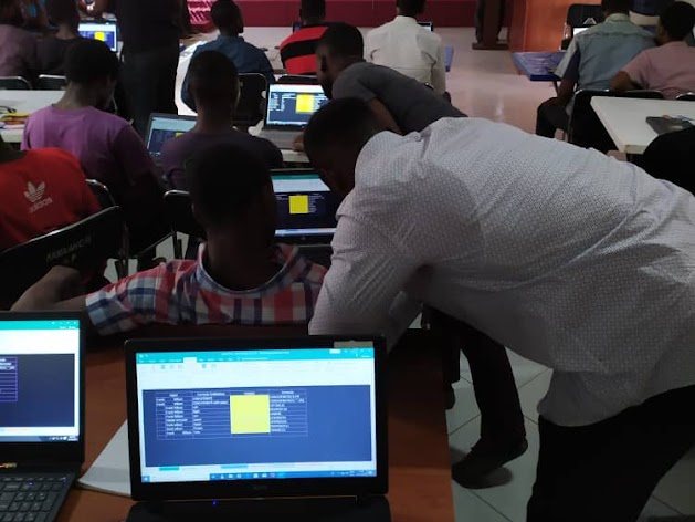 A mentor leaning over to help a learner at a laptop during a classroom coding workshop