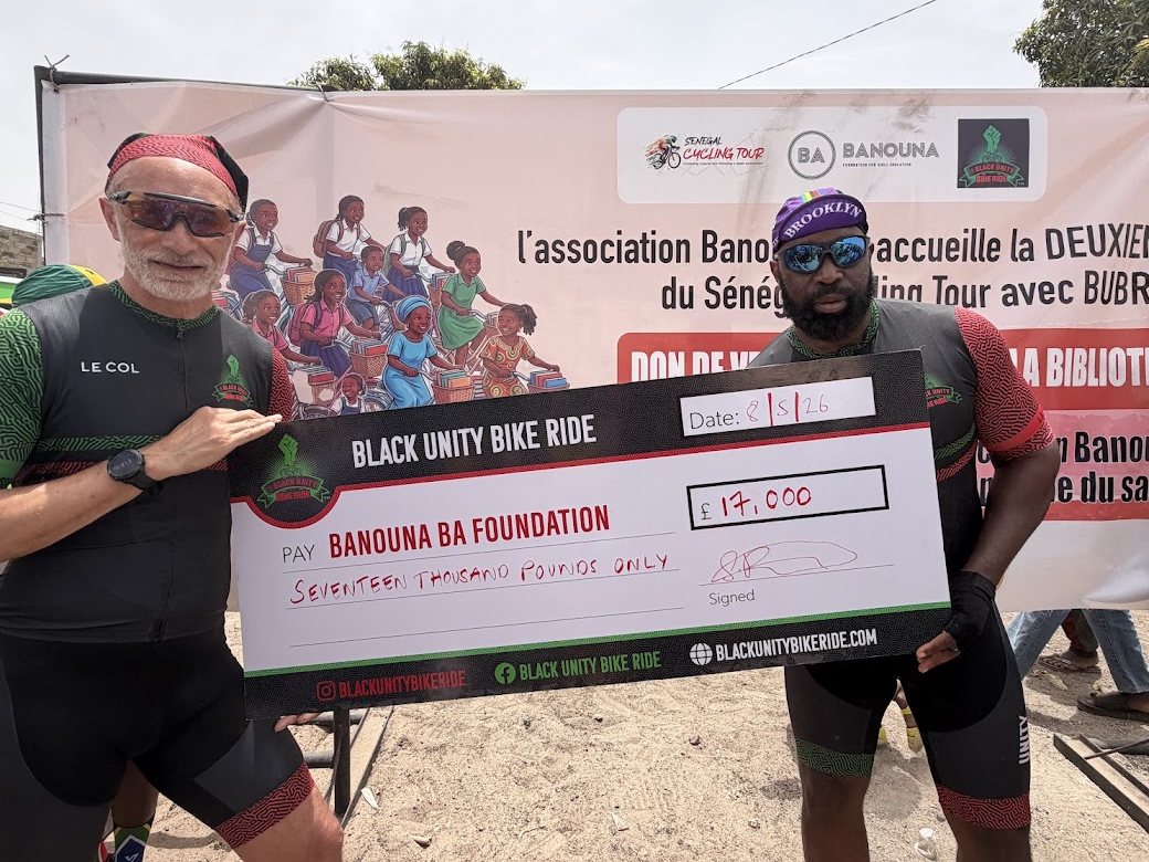 Kenny Akpan in a Brooklyn cycling cap and a fellow Black Unity Bike Ride rider holding a charity cheque in Senegal
