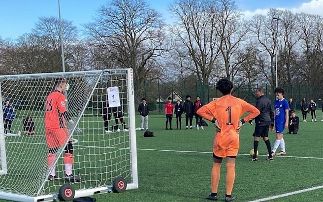 A youth soccer penalty shootout on an artificial pitch, with teams watching from the halfway line