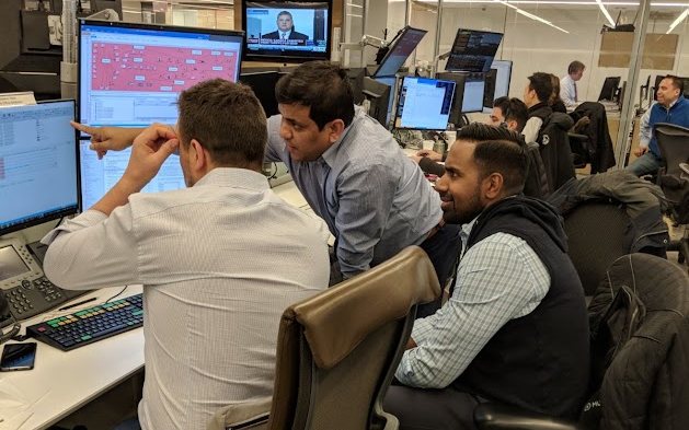 Colleagues gathered around trading-desk monitors, one pointing at data on screen